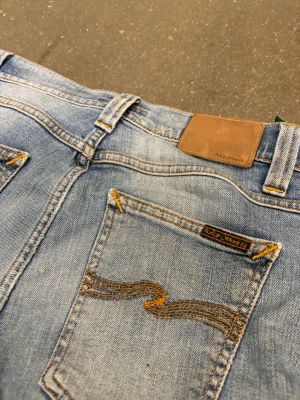 Ljusblå jeans från Nudie Jeans - Säljer ett par ljusblå jeans från Nudie Jeans med klassisk femficksmodell och snygga slitningar. Jeansen har kontrastsömmar, normal midja och go passform. Materialet är mjuk denim i bomull och bakfickorna har Nudie Jeans karakteristiska brodyr. Storlek 30:30