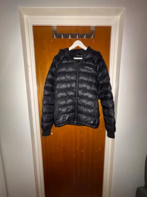 Svart pufferjacka med huva (peak performance) - Säljer en svart pufferjacka med huva och dragkedja framtill. Jackan har en quiltad design och två sidofickor. Perfekt för kalla dagar och ger en sportig look. Materialet ser ut att vara syntetiskt och jackan är lätt vadderad för extra värme.