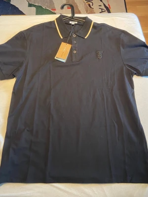 Burberry  - Snygg svart pikétröja med guldfärgade detaljer på kragen och broderad logga på bröstet med märket Burberry . Perfekt för dig som vill ha en stilren och trendig look. Den är 50cm axelbred och 74 cm lång. Fraktar samma dag som den säljs. Den är helt ny. 