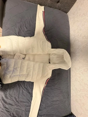 Moncler cardigan  - Snygg vit pufferjacka med huva och ribbade partier. Jackan har långa ärmar med röd, vit och blå rand längs ärmarna för en sportig look. Dragkedja framtill och quiltad framsida. Perfekt för kyligare dagar. Oanvänd!!, scann på loggan fungerar.
