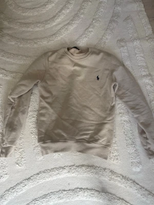 Beige sweatshirt från Ralph Lauren - Säljer en stilren beige sweatshirt från Ralph Lauren med klassisk rund halsringning och broderad logga på bröstet. Tröjan har långa ärmar och ribbade muddar vid ärmslut och nederkant. Perfekt för en avslappnad och snygg look.