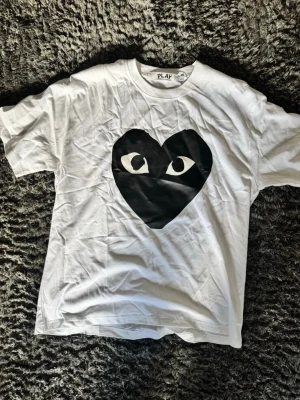Vit CDG T-shirt med svart hjärta - Vit CDG T-shirt med ett stort svart hjärta och ögon på bröstet. Klassisk passform och rund halsringning. Tillverkad i mjuk bomull, perfekt för en avslappnad stil. Enkel men ändå unik design som sticker ut.