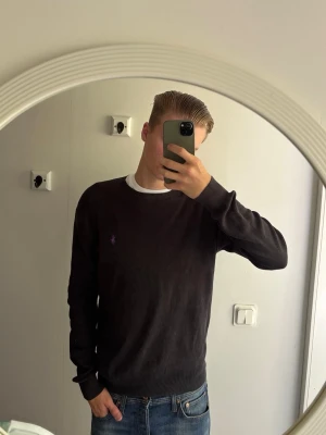Svart tröja från Ralph Lauren - Klassisk svart tröja från Ralph Lauren med rund halsringning och den ikoniska lila broderade loggan på bröstet. Tröjan har långa ärmar och ribbade muddar vid ärmslut och nederkant. Perfekt för en stilren och enkel look. Den är lite paj vid hängaren men kan laga innan köpet!