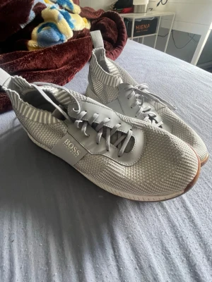 Grå stickade sneakers från BOSS - Snygga grå sneakers från BOSS med stickad ovandel och detaljer i läder. Skorna har vita snören, ribbade kanter och en brun sula med HUGO BOSS-logga framtill. Perfekta för dig som gillar stilrena och moderna sneakers. använt bara i en vecka!!