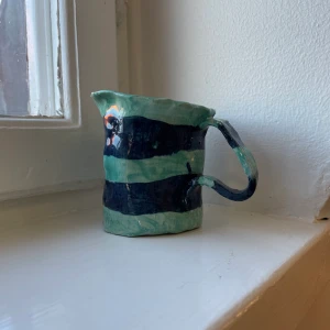 En handgjord grön kopp med blåa ränder  - En handgjord kopp i keramik. Koppen är ljusgrön med mörkblåa ränder medan insidan av koppen är ofärgad. 