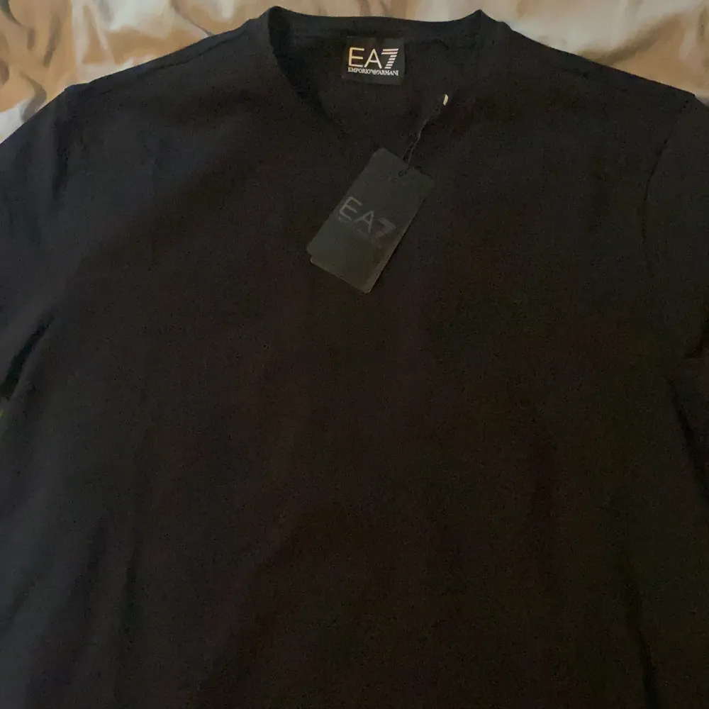 En svart Emporio Armani t-shirt med svart ”EA7 Emporio Armani” logo på bröstet samt vit text på ryggen. Tröjan är aldrig använd och kommer tillsammans med påsen, köpt på NK. Anledning till varför jag säljer är att den ej passade mig storleksmässigt. Urpringspris: 1000kr. T-shirts.