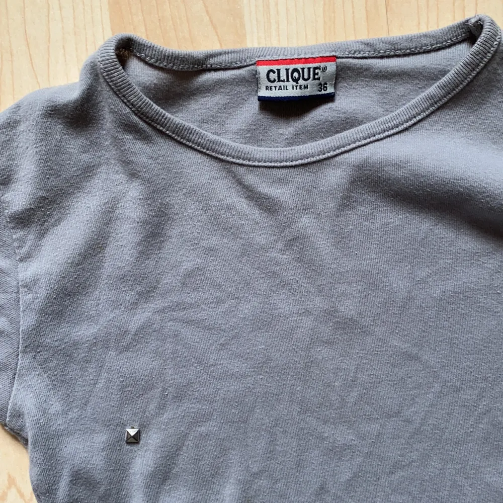 En grå t-shirt i figursmickrande form från CLIQUE. Har endast använts ett fåtal gånger och har en diamant på högra bröstet. Tyvärr, har stygnen gått upp ytterst lite på baksidan men annars är den i utmärkt skick.. T-shirts.