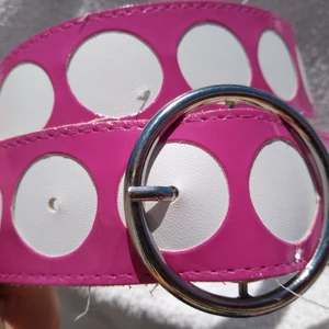 Rosa prickigt bälte i vinyl - Rosa retro bälte med stora vita prickar och cirkelformat silverspänne. Materialet är glansig vinyl. Sparsamt använt. 100 cm långt och4 cm brett. 