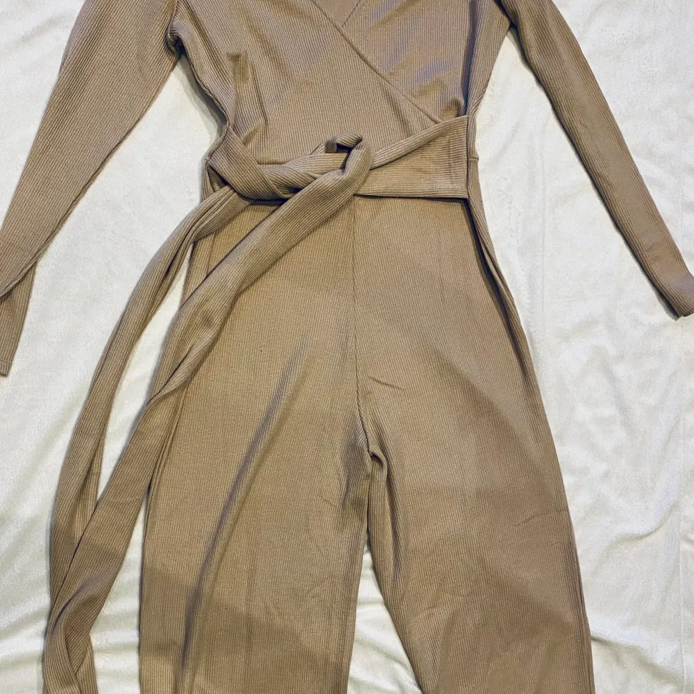 Oanvänd snygg jumpsuit i stickat/ribbat material med knytning i midjan för snygg passform.  Färgen är beige . Stickat.