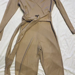 Oanvänd Jump suit  Stl 38 - Oanvänd snygg jumpsuit i stickat/ribbat material med knytning i midjan för snygg passform.  Färgen är beige 
