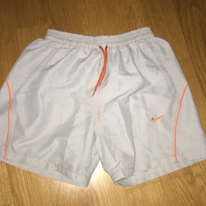 Nike badbyxor  - Vita, lite kortare badbyxor för herrar. Supersnygga med orange logga och skönt material. Storlek M