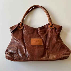 D&G väska - Stor brun Dolce & Gabbana väska, förmodligen inte äkta. Har mågra defekter som syns på bild men inget som störs. Längd: ca 45 cm. Bredd: ca 28 cm. Djup: 11 cm 💕