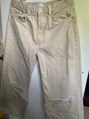 Beige Bershka Jeans - Beige bershka jeans som är i bra skick, knappen kan vara lite trög att sätta på men annars är den jättebra! Storleken är EU 38 och är en wide leg. Jeansen har två hål vid knäna. Jeansen är också   high waist.