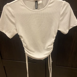 Vit ribbad t-shirt med snören man kan dra åt - Skick är bra, nästan aldrig använd. Enligt mig framhävs brösten i tröjan. Passar till vilka byxor som helst egentligen då den är vit och basic. 