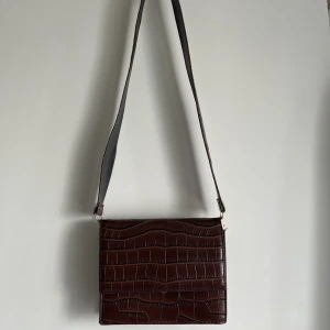 Liten väska - Liten, väldigt praktisk och snygg brun axelremsväska med brett axelband, tvåfärgat (brunt på ena sidan och svart på andra). Mått: 18x15 cm. Axelbandet är borttagbart och totalt ca 90 cm långt, väldigt bra längd för både cross-body och att ha över axeln. 