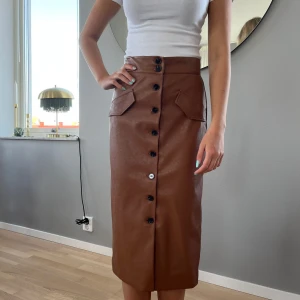 Kjol i skinnimitation  - Brun kjol i skinnimitation med knappar fram. Vadlång. Jättefin till stickad tröja i höst! Lite liten i strl skulle jag säga. Highwaist. 