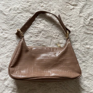 Beige väska i krokodil mönster och guldiga detaljer  - Brun/beige färg Har guldiga detaljer. Är 27cm bred på det bredaste stället och 16cm hög på det högsta. Har stadga i sig så den inte ”sackar” ihop även om de inte är något i väskan 