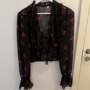 Blus med knytning  - En vacker och tunn blus i rött och svart med knytning vid brösten. Det är volanger längs med blusen och även vid händerna. 