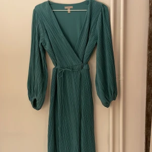 Omlottklänning - En superfin vadlång omlottklänning med plisserad festlig material från H&M. Långa ballongärmar och v-ringad. Storlek M. Färgen är turkos, en blandning mellan grön och blå. Använt två gånger, nästan som ny.