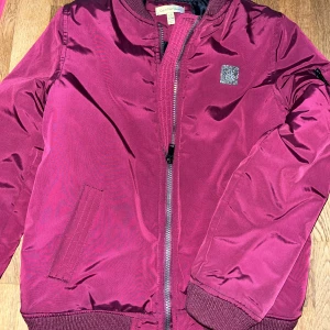 Calvin Klein  - Jacka från Calvin Klein lätt fodrad.  Vinröd, blank i tyget. Jackan är i nyskick. Amerikansk storlek 12/14 motsvarar cirka 152/158 cl. 