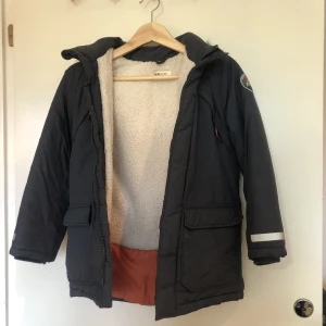 Gråblå vinterjacka för barn - Fluffig vinterjacka i stl 134 från H&M. Med två större och två mindre fickor på framsidan, avtagbar luva med pälskrage och reflexer. Fodret är något slitet i nederkant. 