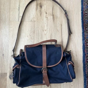 Axelbandsväska från Massimo Dutti - Fin axelbandsväska med detaljer från Massimo Dutti, finns innerfickor och stängs med magnetknapp. Mörkblå med brunt läder, axelbandet går att justera. Bra skick!