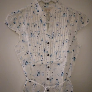 Feminin vintage look kortärmad blus. Vit / blå. Blommig. BikBok. Strlk: XXS-XS.  - Feminin vintage look kortärmad blus. Vit / blå. Blommig. Från BikBok. Strlk: XXS-XS. Bomull. Stängs med tryckknappar och har knytband i midjan. Ok skick, se bilder. Inget trasigt.