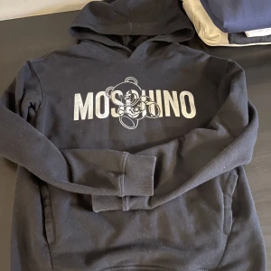 Moschino-tröja - Säljer en fin hoodie från Moschino i storlek 152, inköpt på Kids Brand Store december 2022. Svart, inga hål, djur- och rökfritt hem. Nypris 1200kr