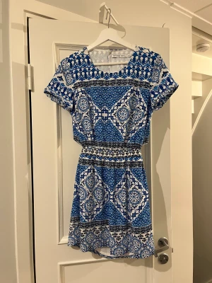 Härlig blå och vit mönstrad klänning  - Klänningen är relativt kort och har en bar rygg öppning. Den har ett resårband som ger klänningen en fin passform. Den har även två fickor på vardera sida. Den har används ett flera gånger och är i mycket bra skick. Den är normal i storleken