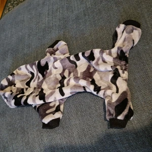 Grå overall till hund  - Kamouflagemönstrad, med huva, knäpps på magen med tryckknappar. Ryggmått ca 35cm, framben ca 12cm, bakben ca 14cm. Väldigt mjukt och lent material. Använd ett fåtal gånger av en nakenhund, så inga pälsrester finns.