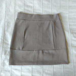 Beige figursydd och elastisk minikjol, storlek 36 - Beige/greige kort kjol. Storlek 36. Elastisk och sitter jättefint på. Resår i midjan. Man kan välja lite hur högt upp i midjan man vill ha den. I fint skick. 97% polyester 3% elastan.