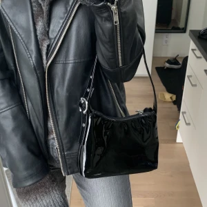 SILFEN ”Ulla bag” väska - Svart väska i glossy vegant läder med silvrig bältesdetalj på väskbandet från SILFEN, som heter ”Ulla bag”, nypris 749kr. Stängs med dragkedja. Axelväska. Jättefint skick.