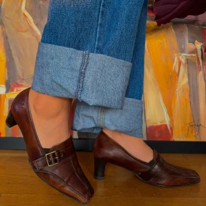 Bruna skinnskor med klack och spänne - Säljer ett par eleganta bruna skinnskor från Caprice. De har en låg klack och ett dekorativt spänne på ovansidan. Skorna har en spetsig tå och är i storlek 38.5. Perfekta för både vardag och fest!