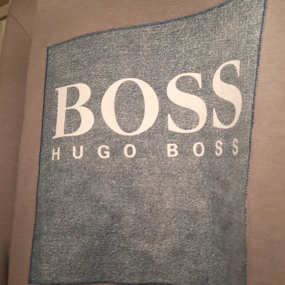 Säljer en stilren grå sweatshirt från Hugo Boss. Den har en stor, blå fyrkant med BOSS-loggan i vitt på framsidan. Tröjan är långärmad och har en normal passform, perfekt för både vardag och avslappnade tillfällen. Materialet känns mjukt och bekvämt.. Tröjor & Koftor.