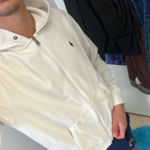 Vit hoodie från Ralph Lauren - Säljer en stilren vit hoodie från Ralph Lauren. Den har en dragkedja framtill och en klassisk logga på bröstet. Perfekt för en avslappnad och snygg look. Passar till alla tillfällen!