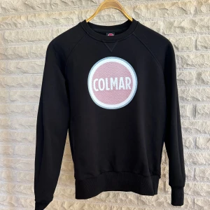 Svart sweatshirt från Colmar - Säljer en snygg svart sweatshirt från Colmar med deras ikoniska logga i vitt och rosa på bröstet. Tröjan är långärmad och har en bekväm passform, perfekt för både vardag och chill. Den är i bra skick och redo för en ny ägare!