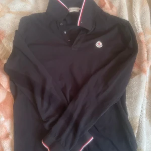 Svart pikétröja från Moncler - Säljer en snygg svart pikétröja från Moncler. Den har långa ärmar och en klassisk krage med vita och röda detaljer. Perfekt för både vardag och lite finare tillfällen. Tröjan är i bra skick och kommer i originalförpackning.