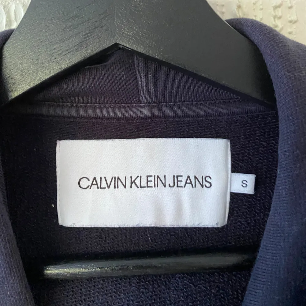 Säljer en snygg marinblå hoodie från Calvin Klein Jeans. Den har en CK-logga i vitt, blått och rött på bröstet och texten 'Calvin Klein Jeans' längs ena ärmen. Perfekt för höst och vinter med sin mysiga passform och känguruficka framtill. Huvan har dragsko för justering. Bra skick. Hoodies.