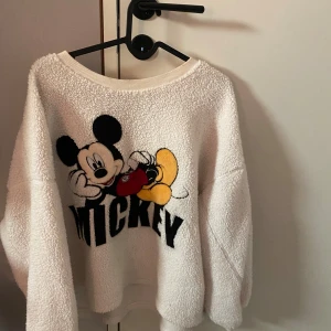 Vit fluffig tröja med Musse Pigg från H&M - Säljer en supermysig vit fluffig tröja från H&M med ett stort tryck av Musse Pigg på framsidan. Tröjan har en lös passform och är perfekt för kalla dagar. Den är långärmad och har en ribbad kant vid halsen. Perfekt för att mysa i hemma eller för en avslappnad dag ute! Sen även ett par mysiga byxor till, något smink på kragen på tröjan, använt fåtal gånger. Hör gärna av er vid frågor eller fler bilder💕