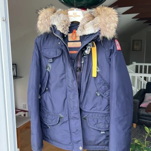 Blå vinterjacka från Parajumpers - Säljer en snygg blå vinterjacka från Parajumpers, perfekt för kalla dagar. Jackan har en fluffig pälsfodrad huva och flera praktiska fickor. Den är i slim fit och har coola detaljer som en gul rem vid kragen och en patch på ärmen. Mycket fint skick!