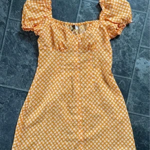 Gul blommig klänning - Säljer en superfin orange klänning med vita blommor. Den har puffiga kortärmar och söta knappar framtill längst klänningen. Perfekt för sommardagar och fester! 💛