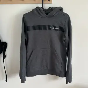 Säljer en snygg grå hoodie från Sail Racing. Den har en svart rand över bröstet med loggan tryckt på och texten 'SAIL RACING' på huvan. Perfekt för en casual look och superbekväm med sin mjuka insida. Passar perfekt till höst och vår!