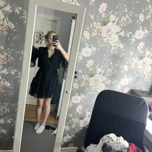 Svart klänning med spetsdetaljer - Snygg svart klänning med spetsdetaljer och v-ringad design. Den har långa ärmar och en kort längd som ger en elegant look. Perfekt för olika tillfällen.