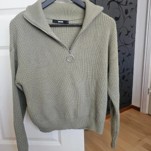 Olivgrön stickad tröja från Bik Bok - Säljer en olivgrön stickad tröja från Bik Bok med dragkedja och hög krage. Tröjan har långa ärmar och en avslappnad passform, perfekt för kyligare dagar. Den är både stilren och bekväm.