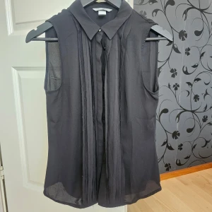 Svart ärmlös blus från H&M - Snygg svart ärmlös blus från H&M med knappar framtill och klassisk krage. Perfekt för en stilren look. Blusen har en lätt transparent design som ger en elegant touch.