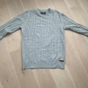 Grå stickad tröja från Bondelid - Säljer en stilren grå stickad tröja från Bondelid. Tröjan har ett klassiskt kabelstickat mönster och rund halsringning. Perfekt för kyligare dagar.