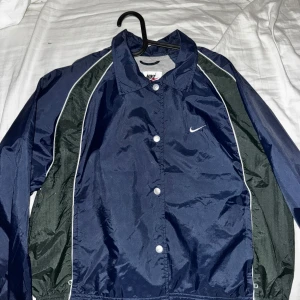 Blå vindjacka från Nike - Säljer en blå vindjacka från Nike med vita och svarta detaljer. Jackan har knappar framtill och en klassisk Nike-logga på bröstet. Perfekt för sportiga aktiviteter eller som en lätt ytterjacka.