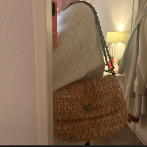 Beige axelväska med kedja - Snygg beige axelväska med kedja och ett klassiskt rutigt mönster. Väskan har en elegant design med en vridknapp framtill. Perfekt för att ge en stilfull touch till din outfit.