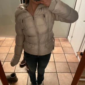 Beige pufferjacka med dragkedja - Säljer en snygg beige pufferjacka med dragkedja och huva. Jackan har långa ärmar och ribbade muddar vid ärmslut. Perfekt för kyligare dagar och även i skidbacken! Storlek 44 men passar som S/M. 