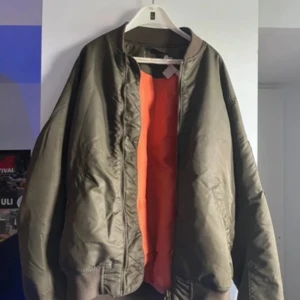 Olivgrön bomberjacka från H&M - Säljer en olivgrön bomberjacka från H&M med orange foder. Jackan har dragkedja och ribbade muddar vid ärmslut och nederkant. Perfekt för en cool och avslappnad stil.